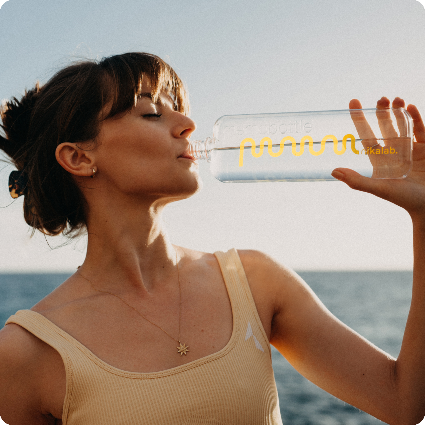 Kobieta w topie w odcieniu nude pije wodę z butelki nikalab limited edition memobottle™ z żółtym logo na tle morza – świadome nawodnienie w stylu nikalab.