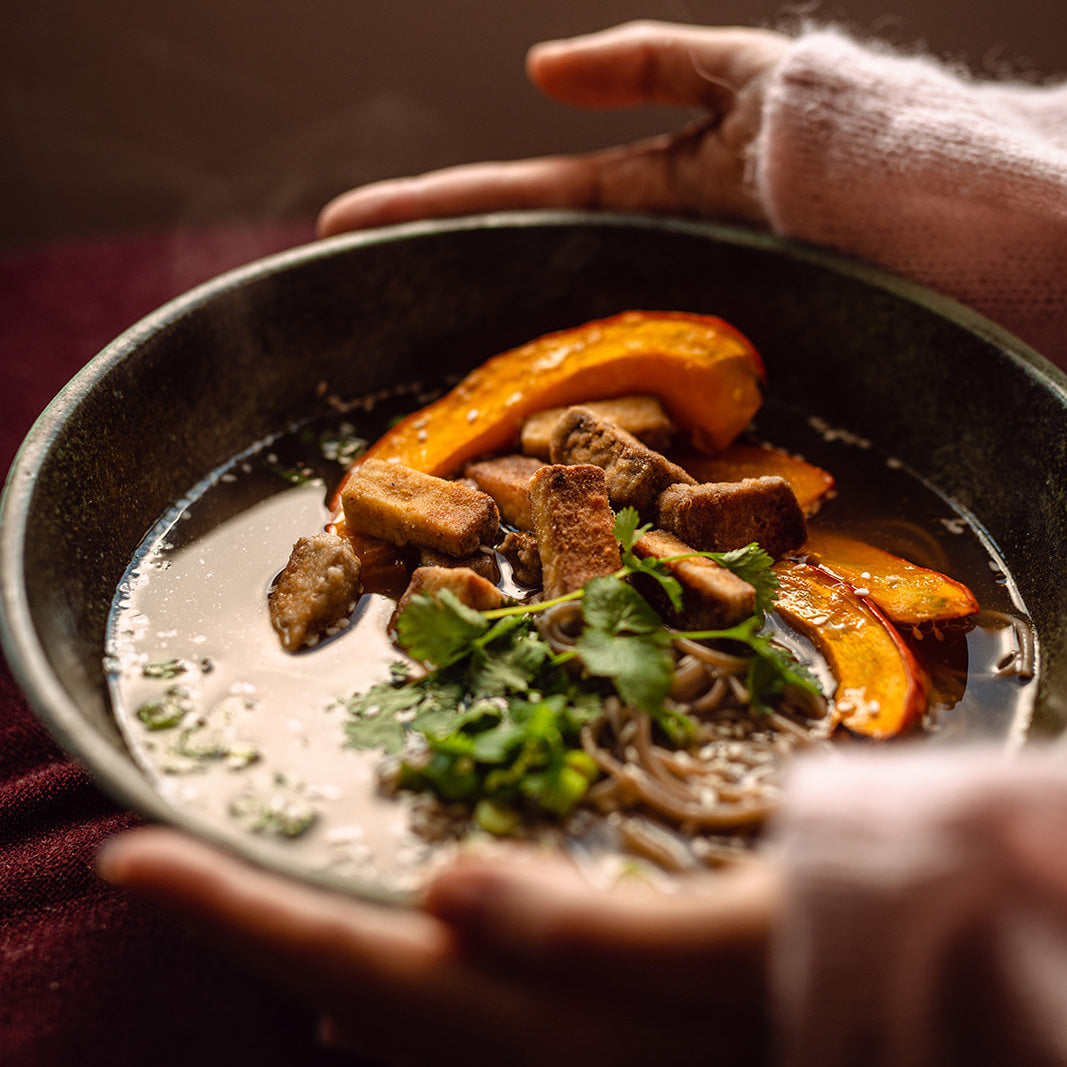 Parująca miska roślinnego ramenu z makaronem gryczanym, smażonym tofu, pieczoną dynią i świeżą kolendrą, podawana w ceramicznej misce – trzymana w dłoniach w swetrze na tle ciepłego światła.
