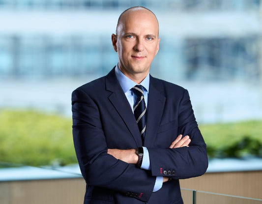 Andrzej Kozłowski w garniturze i pod krawatem, z założonymi rękami, pozuje na tle nowoczesnego biurowca – oficjalny portret biznesowy prezesa.