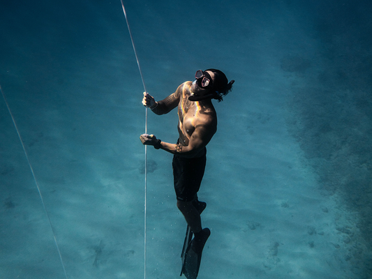 Freediver w masce i płetwach wynurza się wzdłuż liny w turkusowej wodzie, z piaszczystym dnem w tle.