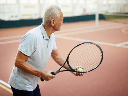 Starszy mężczyzna w jasnej koszulce przygotowuje się do serwisu na krytym korcie tenisowym