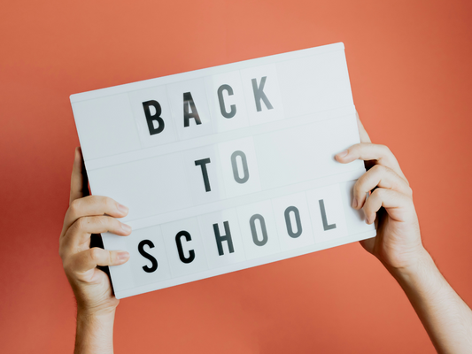 Grafika z napisem Back to School na białej tablicy trzymanej w dłoniach na tle koloru pomarańczowego, symbolizująca powrót we wrześniu do szkoły i zdrowej rutyny.