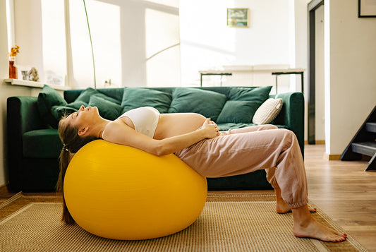 Kobieta w ciąży wykonuje ćwiczenia na żółtej piłce w pokoju, w którym stoi zielona kanapa.
