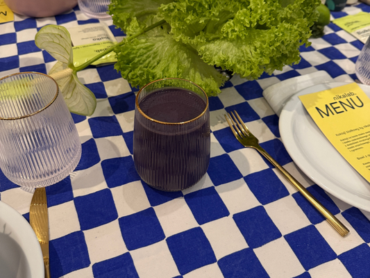 Szklanka czekoladowego koktajlu białkowego z jagodami na stole w niebiesko-białą kratę, obok talerz z kartą menu nikalab i dekoracja z sałaty.