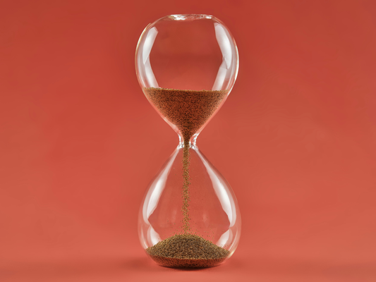 Szklana klepsydra z przesypującym się piaskiem na pomarańczowym tle – symbol upływu czasu i spokojnego podejścia do nudy.