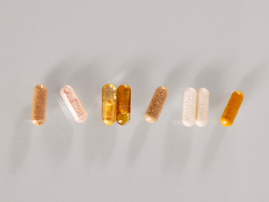 Różne kapsułki suplementów diety w odcieniach bieli i bursztynu ułożone w rzędzie na jasnym tle.