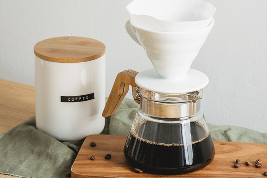 Na blacie kuchennym stoi zaparzona metoda alternatywną kawa. Kawa jest w dzbanku, na którym stoi drip. Dookoła porozrzucane są ziarna kawy 100% arabica.