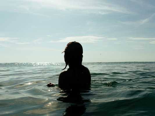 Sylwetka osoby w czapce stojącej w morzu o zachodzie słońca – ilustracja do artykułu o wpływie kontaktu z wodą na stres, sen i koncentrację.