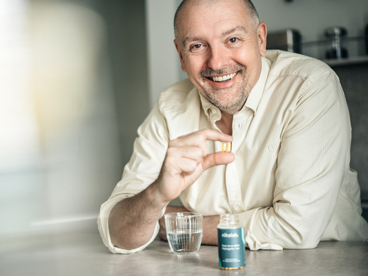 Uśmiechnięta osoba trzyma kapsułkę suplementu nikalab przy szklance wody na kuchennym blacie.