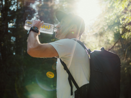 Mężczyzna z plecakiem pije wodę z butelki w lesie; mocne słońce przebija przez korony drzew.