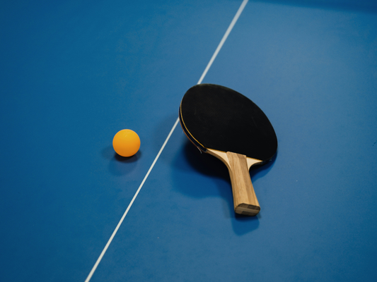 Rakietka do ping-ponga i pomarańczowa piłeczka na niebieskim stole tenisowym przy białej linii.