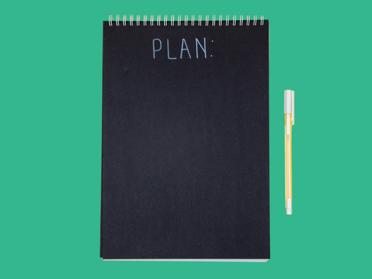 Czarny notes z napisem „PLAN:” i żółty długopis na zielonym tle – symbolizuje planowanie oraz artykuł „Czy nadmiar decyzji odbiera Ci spokój? O odpoczynku bez planu”.