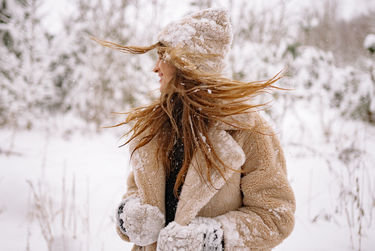 Kobieta w kozuchu i czapce otrzepuje się ze śniegu, dookoła zasnieżone drzewa i las