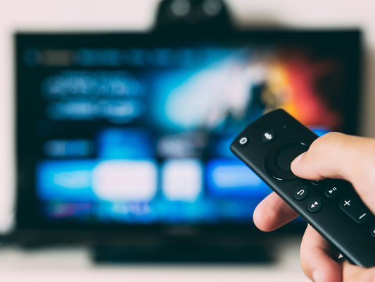 Osoba trzymająca pilot przed telewizorem z włączonym ekranem aplikacji streamingowych, symbolizująca wybór między subskrypcjami takimi jak Netflix, Spotify i usługami zdrowotnymi.