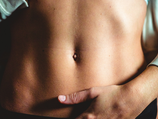 Zbliżenie na brzuch osoby o szczupłej sylwetce, z widocznym pępkiem i dłonią uszczypującą fałd skóry.