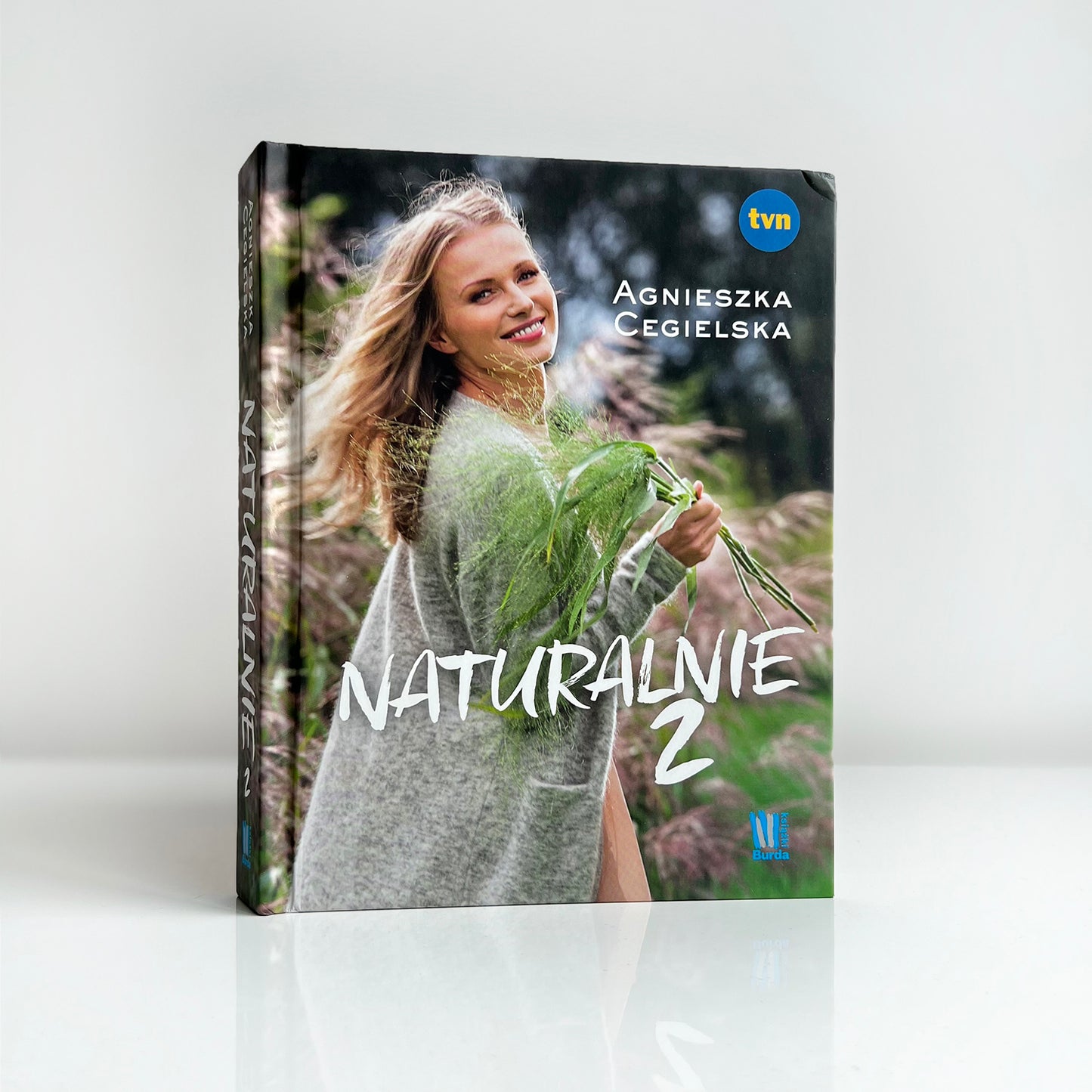 Książka „Naturalnie 2” Agnieszki Cegielskiej – poradnik o zdrowym stylu życia, z okładką przedstawiającą autorkę na tle natury z wiązką traw, logotypami TVN i wydawnictwa Burda.