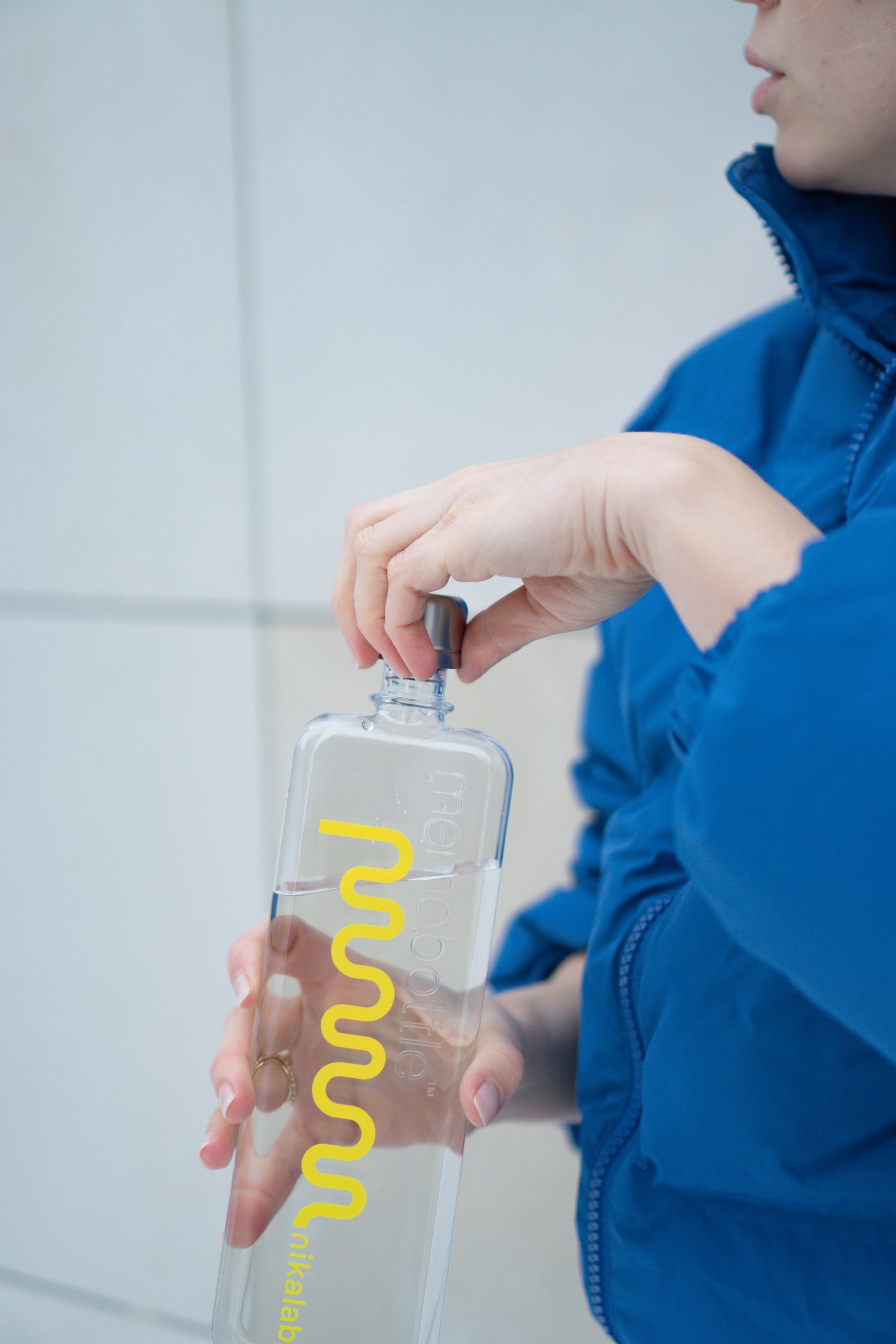 Osoba w niebieskiej kurtce odkręca przezroczystą butelkę memobottle z logo nikalab, częściowo wypełnioną wodą.
