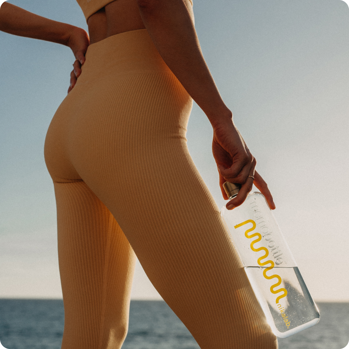 Kobieta w sportowym zestawie w kolorze nude trzyma butelkę nikalab limited edition memobottle™ z żółtym logo, spacerując nad morzem – idealny towarzysz aktywnego stylu życia.