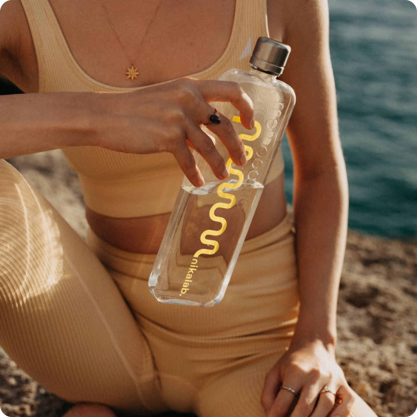 Kobieta w sportowym topie i legginsach nad morzem trzyma butelkę nikalab limited edition memobottle™ z żółtym logo – stylowe nawodnienie w rytmie mindful movement.