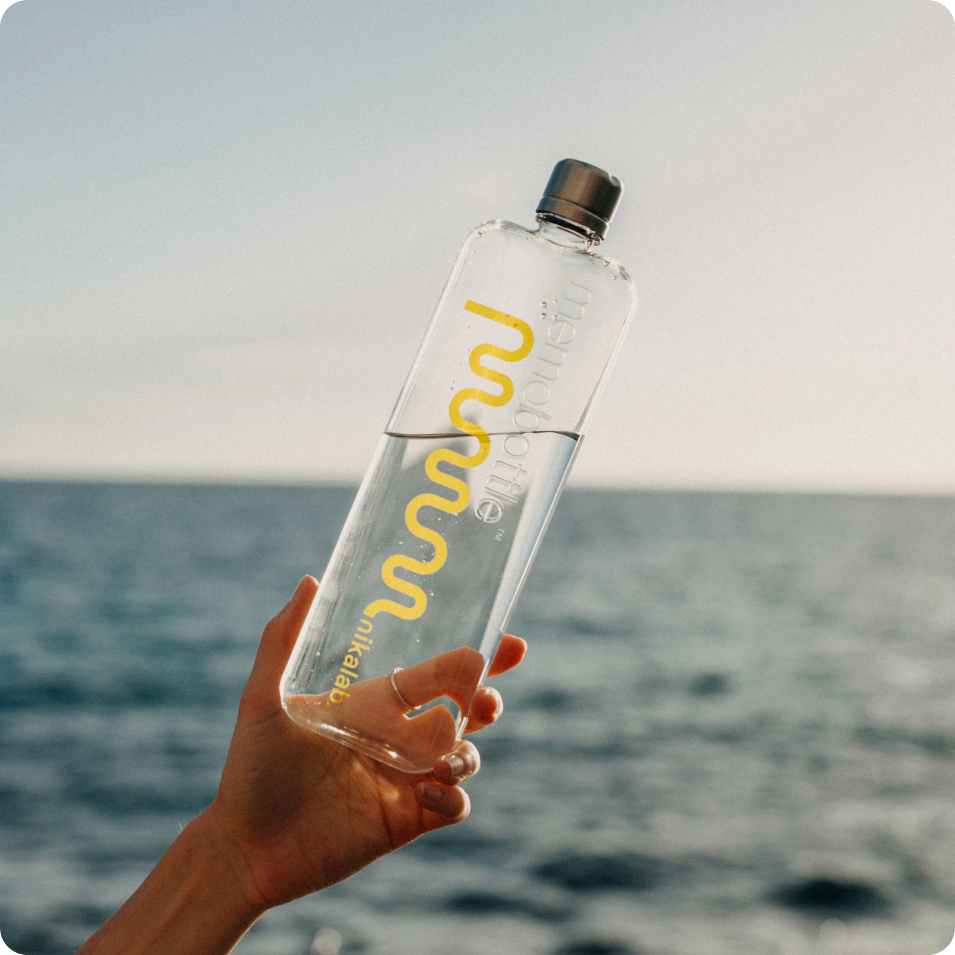Butelka nikalab limited edition memobottle™ z żółtym logo, trzymana w dłoni na tle morza – designerska i ekologiczna alternatywa dla plastiku.