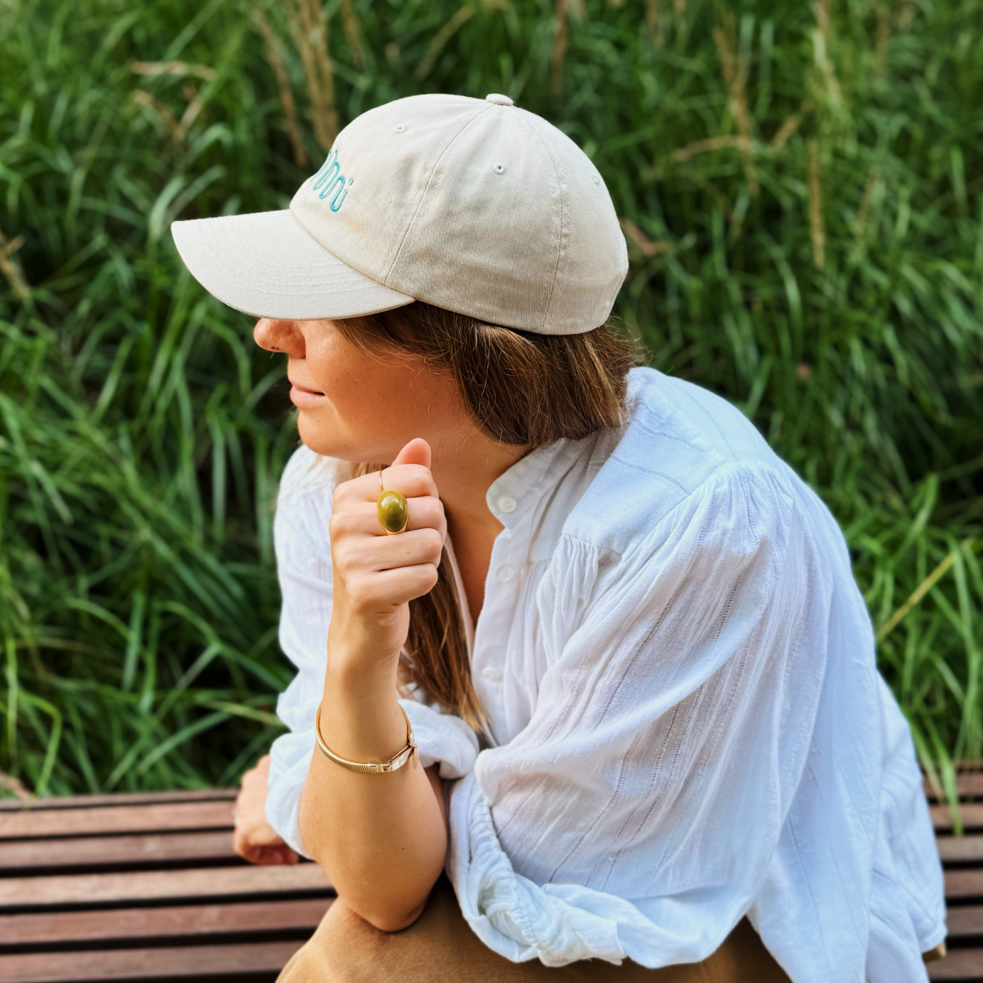 Stylowa kobieta w beżowej czapce z daszkiem nikalab limited edition z miętowym haftem, ubrana w białą koszulę, siedząca na ławce na tle zieleni – idealny look na lato.