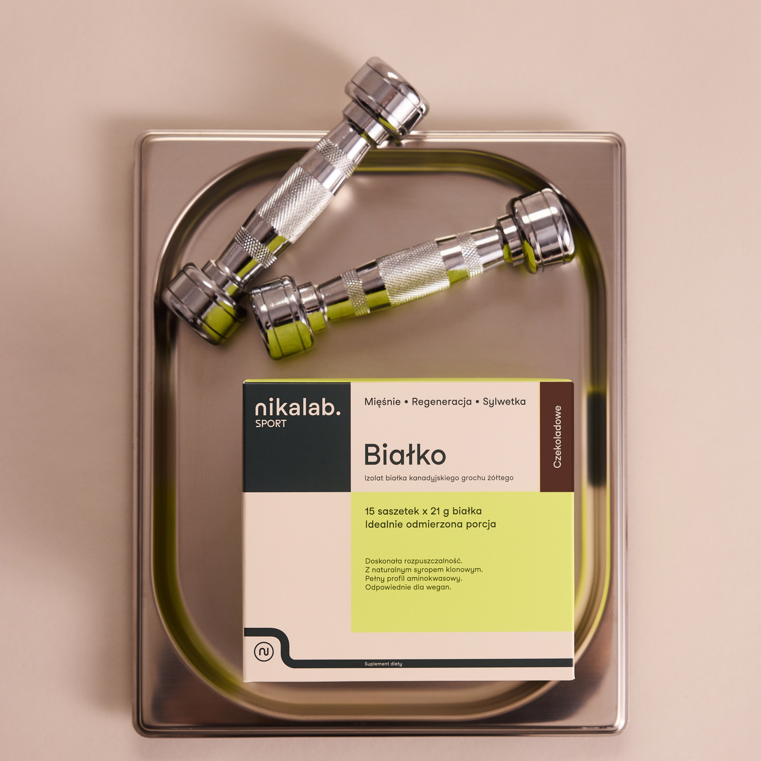 Opakowanie białka nikalab sport w saszetkach o smaku czekoladowym, położone na wadze obok metalowych hantli, na jasnym tle.