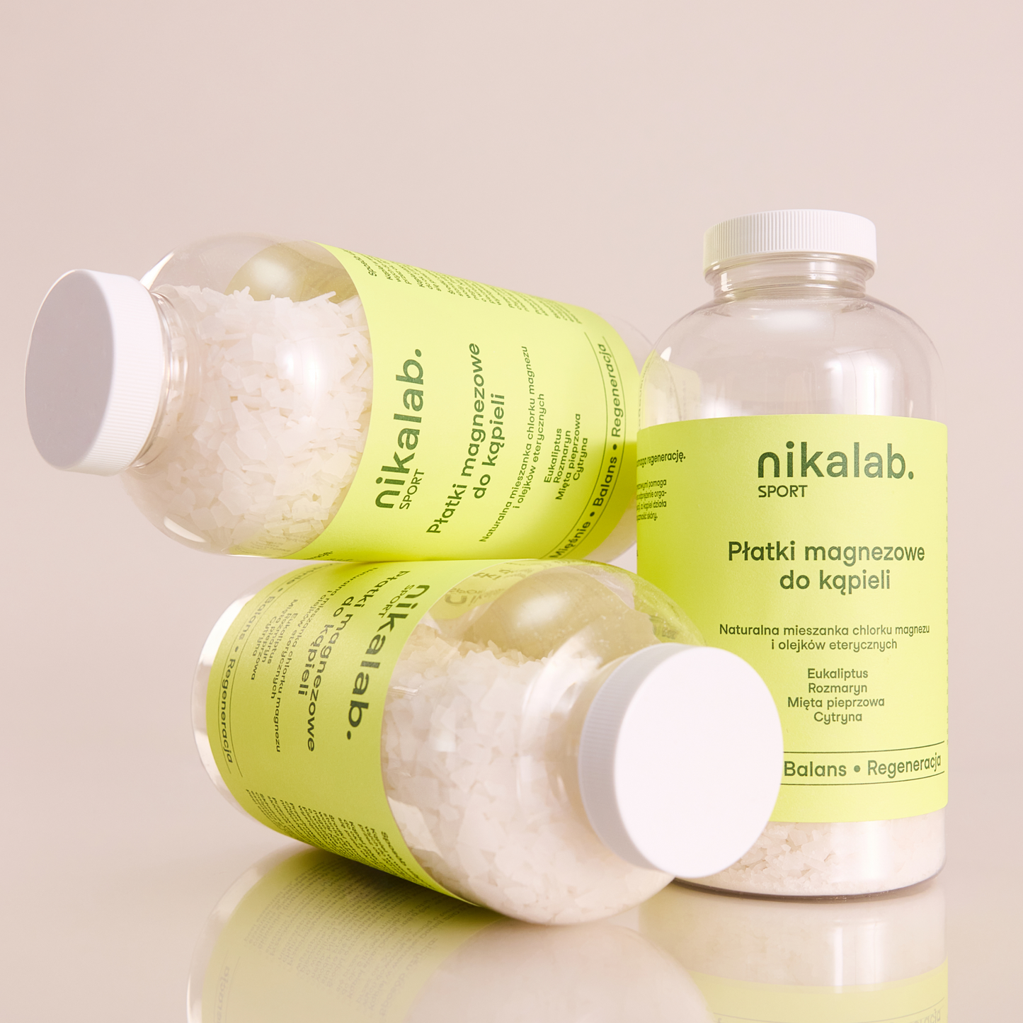 Trzy butelki płatków magnezowych do kąpieli nikalab sport z jasnozieloną etykietą, jedna stojąca i dwie ułożone poziomo, na jasnym tle.