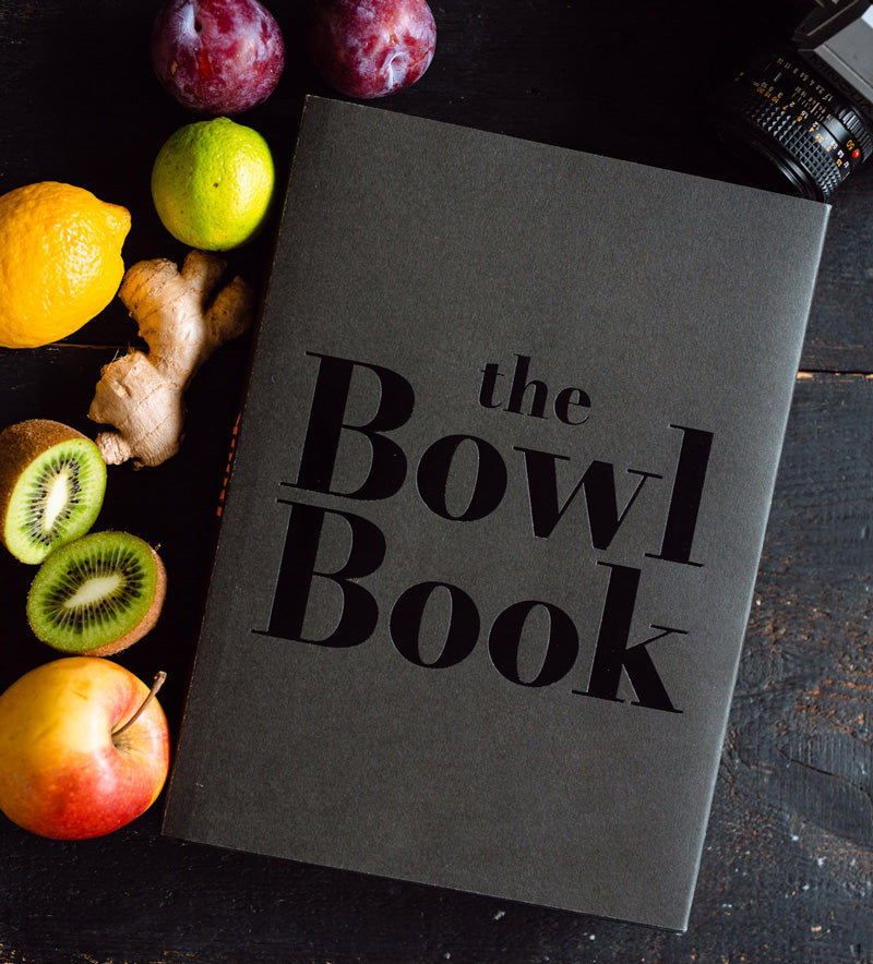 Książka „The Bowl Book” na ciemnym drewnianym stole, otoczona świeżymi owocami (kiwi, jabłko, cytrusy, śliwki) oraz imbirem i aparatem fotograficznym – stylowa kompozycja kulinarna z naturalnym światłem.